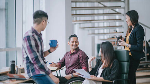 Employees, including one using a wheelchair, discussing work around a desk.