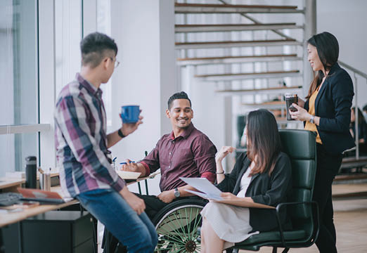 Employees, including one using a wheelchair, discussing work around a desk.