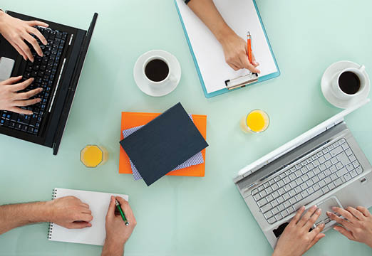 Four people around a table writing in notebooks and typing on computers.