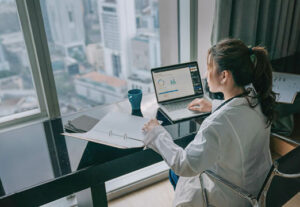 A woman examining a binder of information at a desk with data graphs on a laptop next to her.