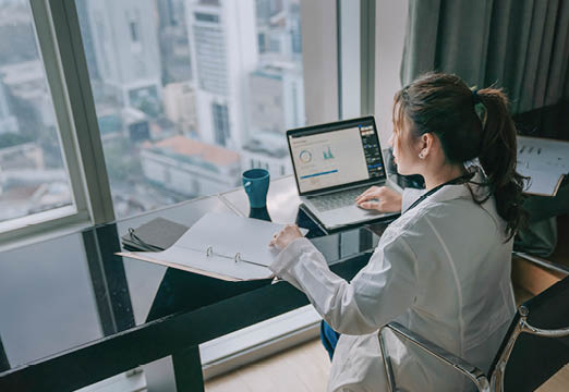 A woman examining a binder of information at a desk with data graphs on a laptop next to her.