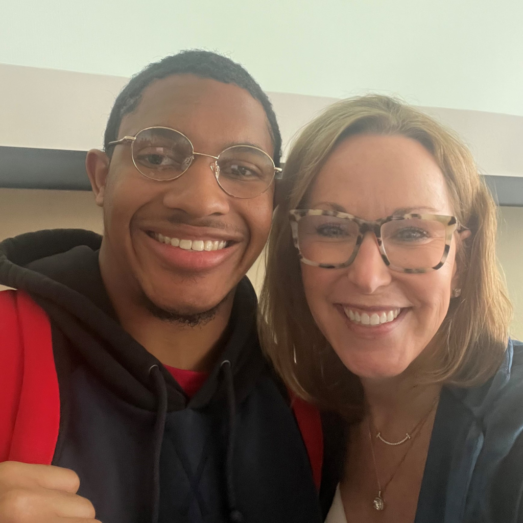 A selfie of two people. One is Keenan an African American young man in a black sweatshirt and glasses and the other is a slightly older white woman in a blazer. They are both smiling at the camera.
