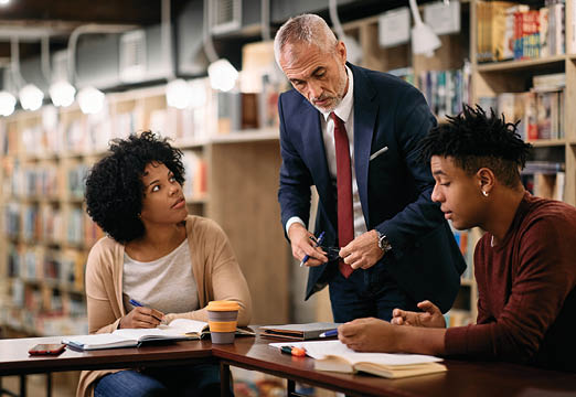 Two students of color talking with a white professor in a library