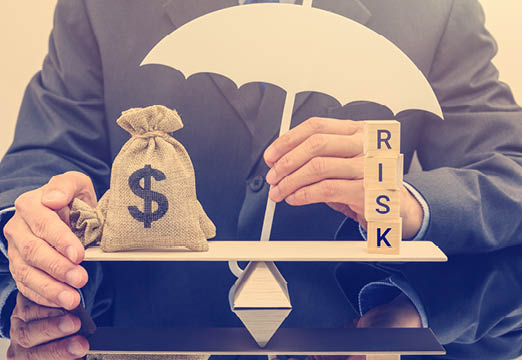A man in a suit balancing a bag of money and blocks that say risk with an umbrella over them.