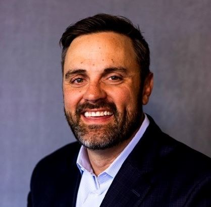 Kenton Miles, a man in a black suit. His button up does not have a tie and the first button is open.