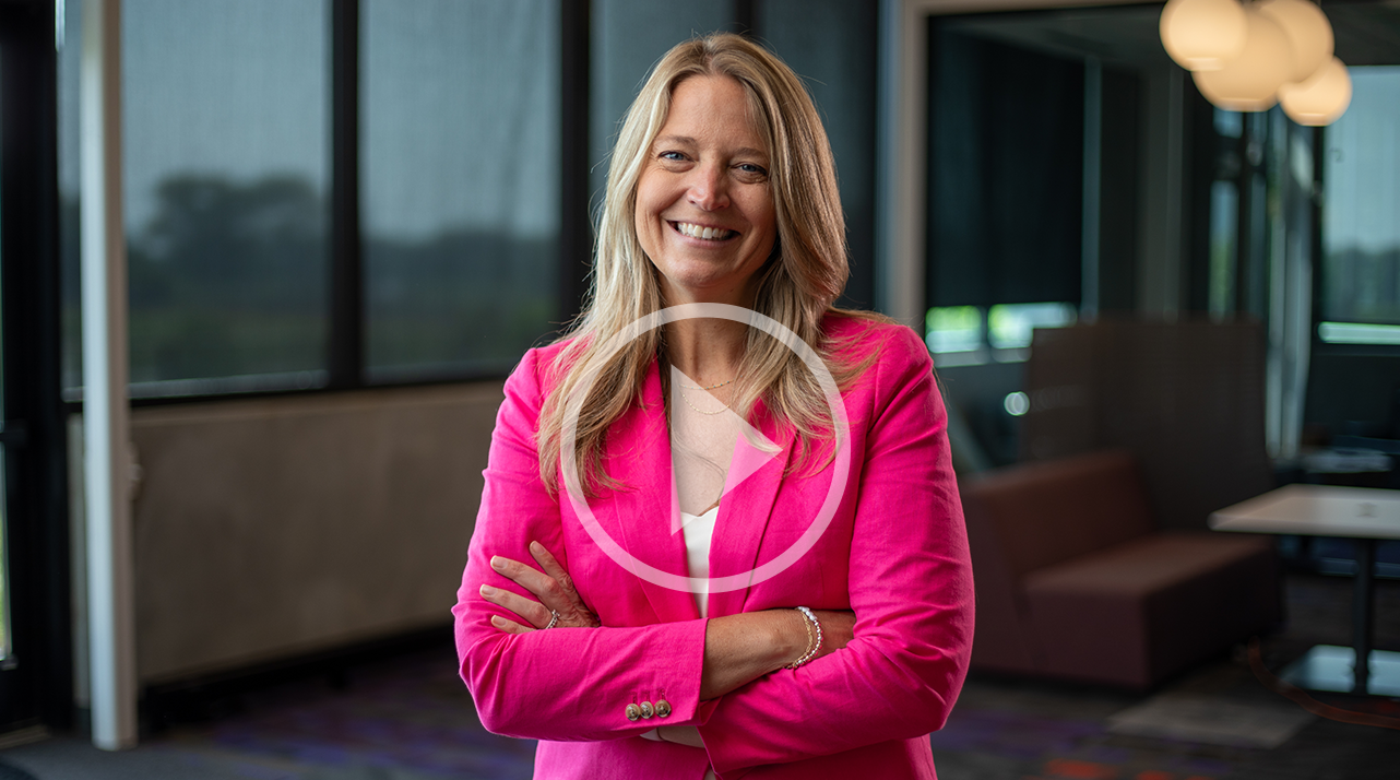 Michelle Weil, a blond woman in a pink blazer smiles for a photo. There is a video play button overlay