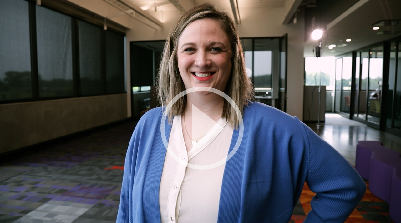 Sara Huston, a blond woman in a blue cardigan, standing for a photo. There is a play button overlay for a video.