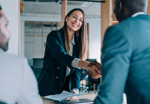 A smiling woman wearing a blazer shaking hands with a man in a suit.