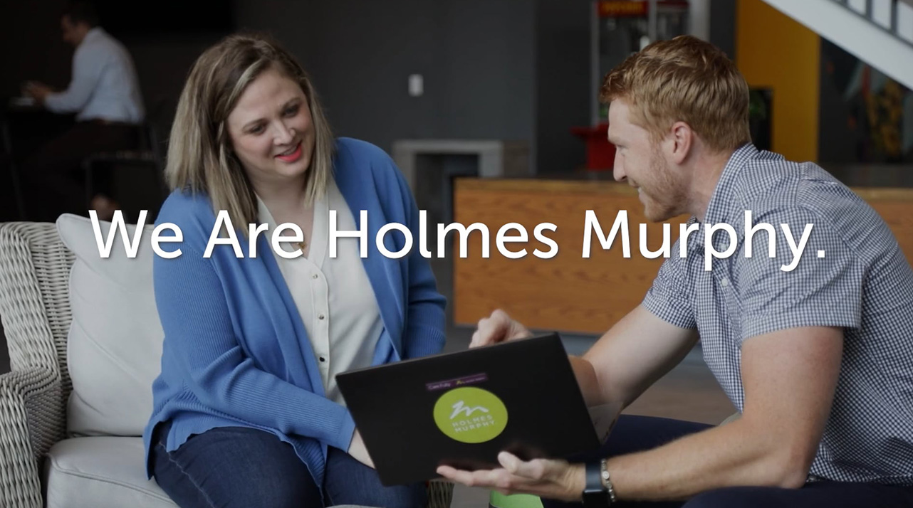 We Are Holmes Murphy: Two employees smiling over a Holmes Murphy laptop.