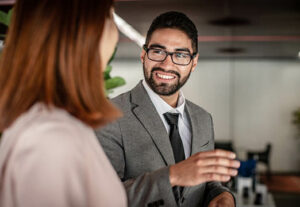 A man in a grey suit chatting with a woman in a pink blouse at work.