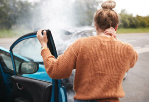 A woman standing in pain with her car that had been in an accident.
