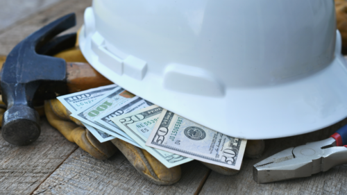 A white construction hat lays on top of cash and a hammer.