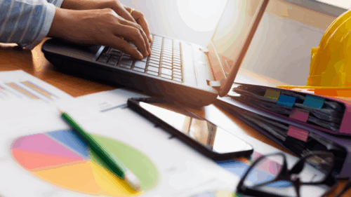 A person with graph documents and binders on their desk types on their laptop
