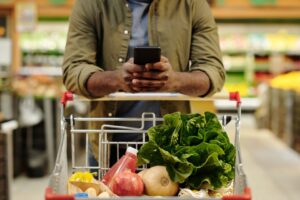 A man with a grocery cart full of produce looks at his phone in a grocery store