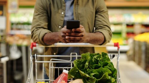 A man with a grocery cart full of produce looks at his phone in a grocery store