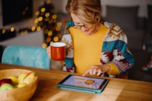A person sips hot cocoa while shopping on a tablet in a cozy and festive living space