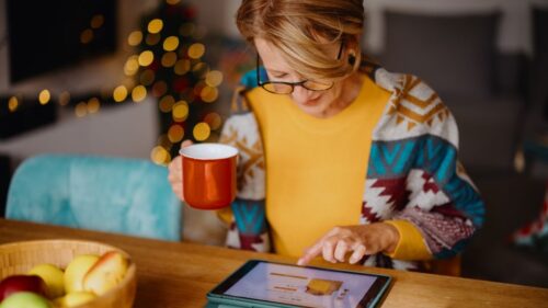 A person sips hot cocoa while shopping on a tablet in a cozy and festive living space
