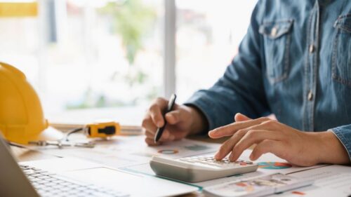 A person uses a calculator while sitting at a desk covered in financial documents and a hard hat and laptop