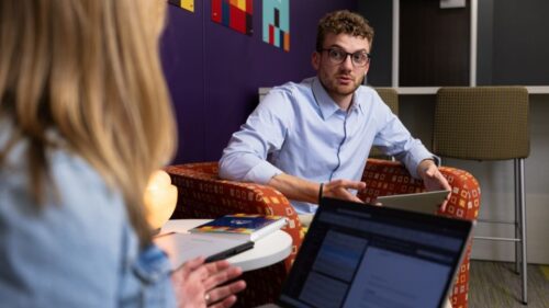 Two employees sit in comfy orange chairs and chat with laptops on their laps