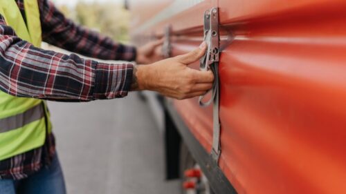 A person wearing a plaid shirt and neon safety vest secures a latch on an orange shipping container.