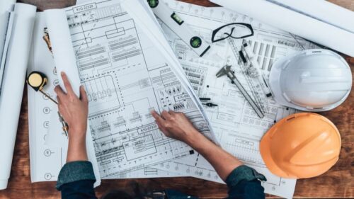 An architect reviews blueprints on a table with the documents, hard hats, and measuring tools.
