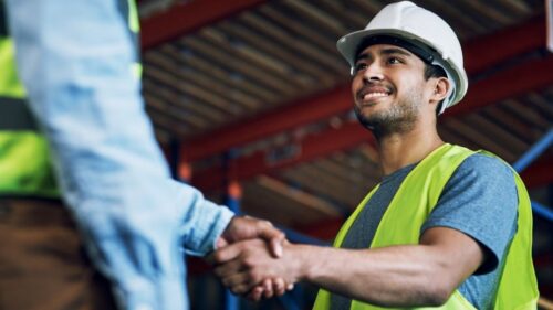 A construction worker wearing a neon safety vest, protective goggles, and a hard hat shakes hands with a supervisor on a job site.