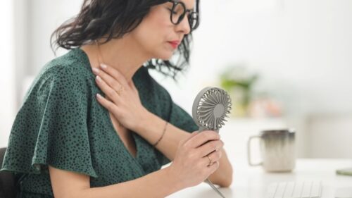 A woman with dark wavy hair in a green dress and black glasses uses a handheld fan during a menopause-induced hot flash.
