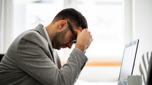 A stressed person sits with their head in their hands in front of a laptop and coffee cup