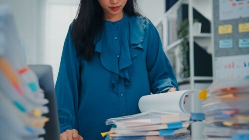 An overwhelmed employee wearing a blue ruffled blouse looks at a stack of papers with multiple tabs and bookmarks.
