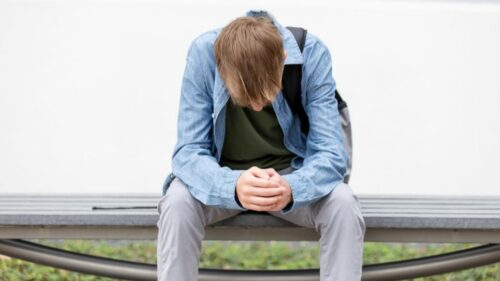 A college student sitting on a bench and looking at the ground.
