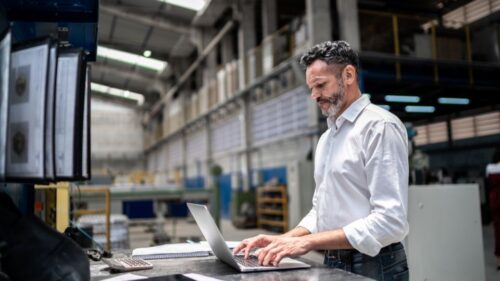 A manufacturing leader typing on a laptop inside of a factory.